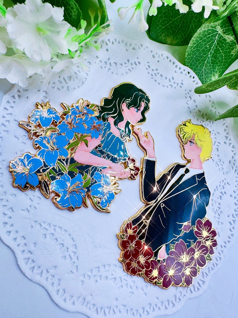 Op de afbeelding: Twee gouden emaille-pins die een stel voorstellen. De vrouw draagt een blauwe jurk en heeft groen haar. Ze is omringd door blauwe bloemen. De man draagt een zwart pak en is omringd door rode bloemen.