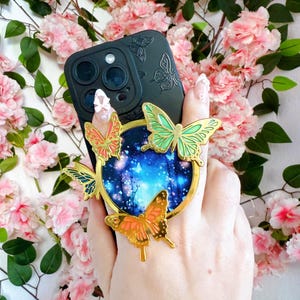 May include: Black phone case with embossed butterfly design. A gold ring with a blue and white night sky scene is adorned with three gold and enamel butterflies in shades of orange and green. The phone case is held against a backdrop of pink flowers.