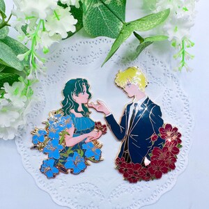 Op de afbeelding: Gouden emaille pinset met een stel in een romantische pose. De vrouw draagt een blauwe jurk met blauwe bloemen en de man draagt een blauw pak met rode bloemen. De pins zijn geplaatst tegen een witte kant achtergrond.