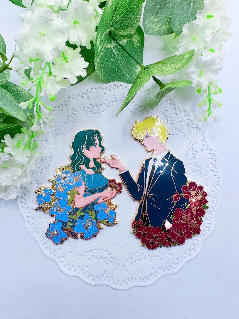 Op de afbeelding: Gouden emaille pinset met een stel in een romantische pose. De vrouw draagt een blauwe jurk met blauwe bloemen en de man draagt een blauw pak met rode bloemen. De pins zijn geplaatst tegen een witte kant achtergrond.