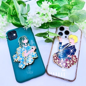 Op de afbeelding: Twee telefoonhoesjes met bevestigde emaille-pins. Het hoesje aan de linkerkant is tealgroen met een blauwe en witte bloemenpin met een vrouw erop. Het hoesje aan de rechterkant is transparant met een rode en gouden bloemenpin met een man erop.