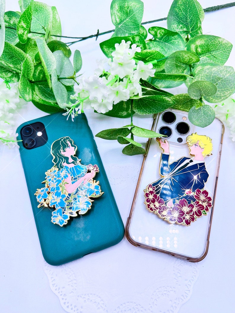 Op de afbeelding: Twee telefoonhoesjes met bevestigde emaille-pins. Het hoesje aan de linkerkant is tealgroen met een blauwe en witte bloemenpin met een vrouw erop. Het hoesje aan de rechterkant is transparant met een rode en gouden bloemenpin met een man erop.