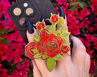 May include: Gold-tone phone grip featuring a bouquet of red roses with green leaves. The phone grip is attached to a black phone case.