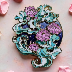 Peut inclure: Épingle décorative représentant des fleurs de lotus roses et des vagues stylisées dans des tons de turquoise, blanc et vert, sur fond bleu foncé. L'épingle a un contour doré et est entourée de pétales de fleurs roses.