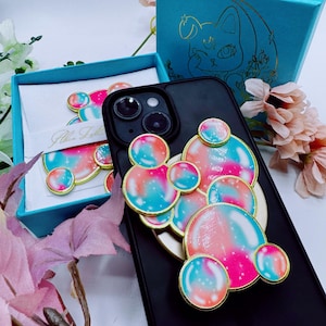 May include: Black phone case adorned with colorful, bubble-shaped embellishments in pink, blue, and gold. A matching set of accessories is displayed in a teal gift box, with a cat illustration on the lid.