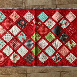 May include: A red quilt with a diamond pattern. The quilt is made up of various fabric squares with different patterns, including floral, polka dot, and plaid. The quilt is edged with a white and blue checked trim.