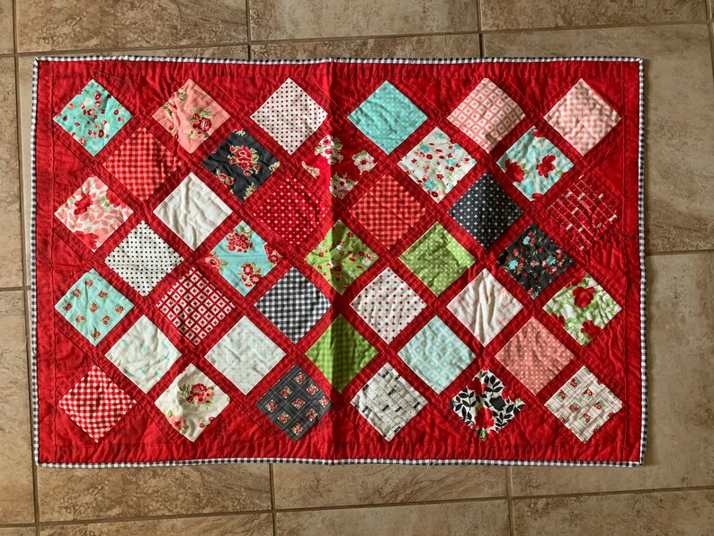 May include: A red quilt with a diamond pattern. The quilt is made up of various fabric squares with different patterns, including floral, polka dot, and plaid. The quilt is edged with a white and blue checked trim.