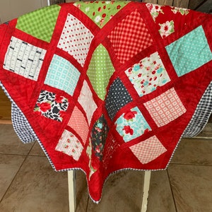 May include: A red quilt with a patchwork design featuring various patterns and colours, including red, green, white, black, and blue. The quilt is folded and draped over a white chair.