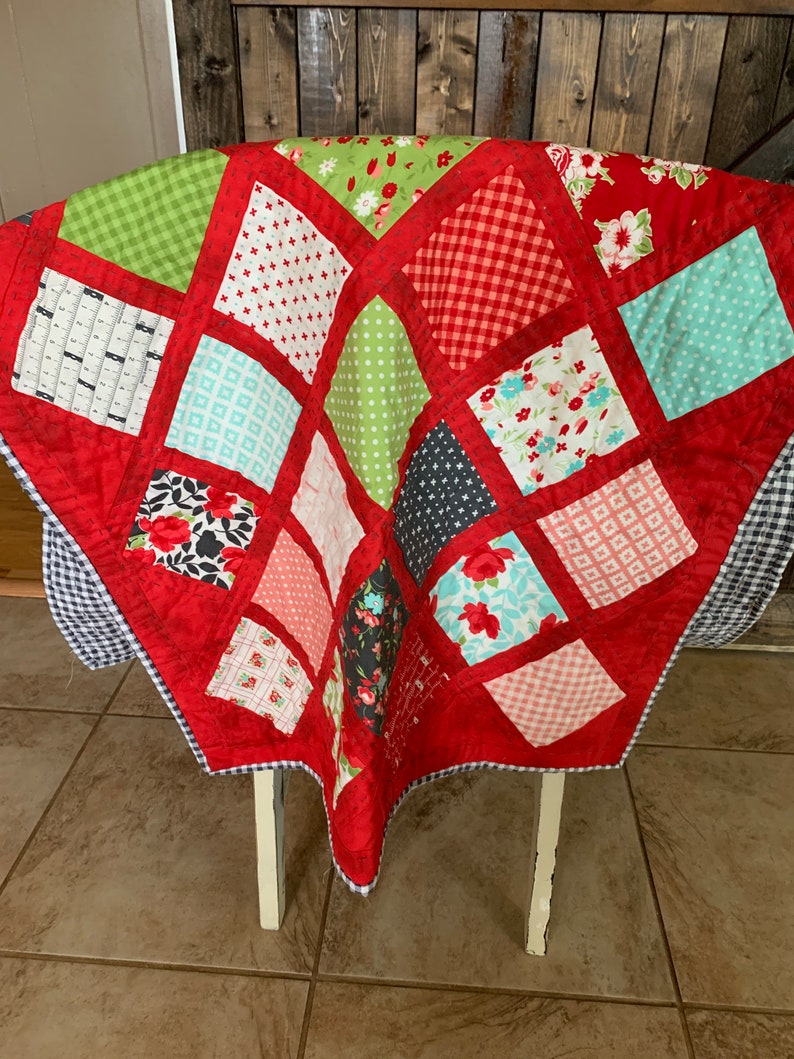 May include: A red quilt with a patchwork design featuring various patterns and colours, including red, green, white, black, and blue. The quilt is folded and draped over a white chair.