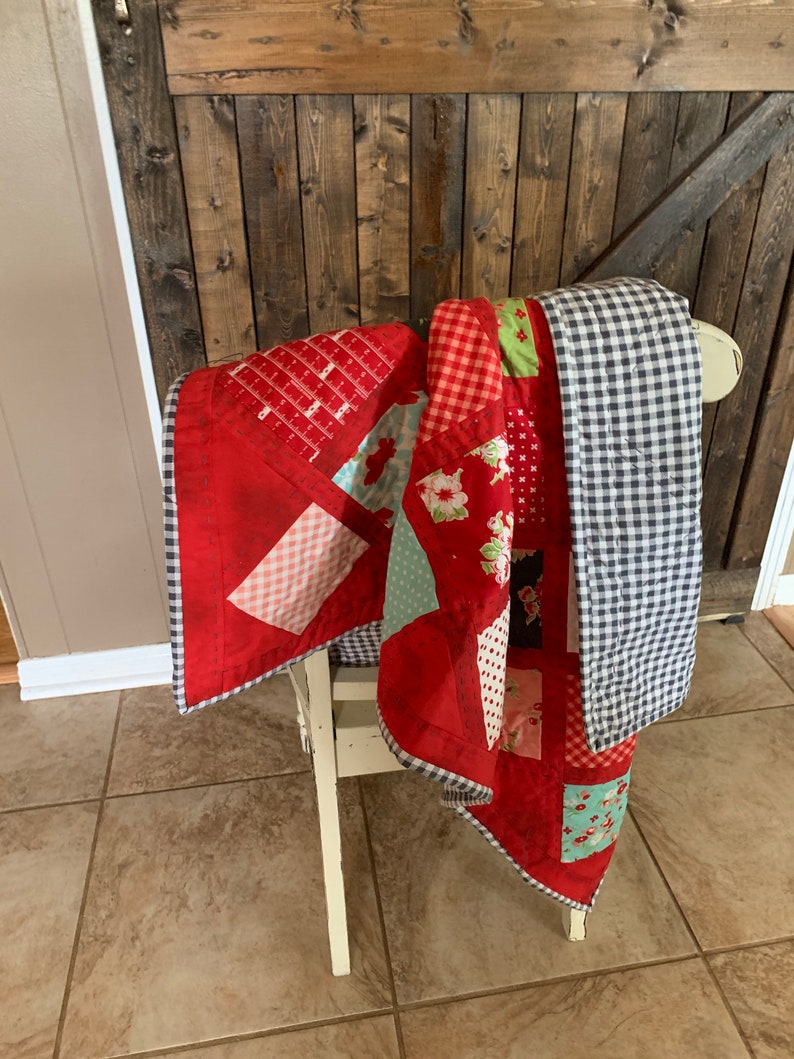 May include: A red, white and blue patchwork quilt with a gingham border is draped over a white chair. The quilt is made of various fabrics, including floral prints, polka dots and geometric patterns.