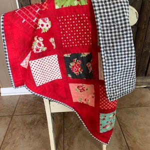 May include: A red, green, black and white patchwork quilt with a black and white gingham backing. The quilt is folded and draped over a white chair.