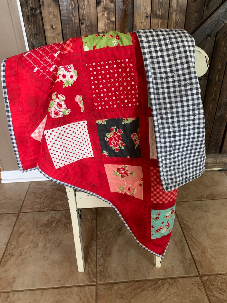 May include: A red, green, black and white patchwork quilt with a black and white gingham backing. The quilt is folded and draped over a white chair.