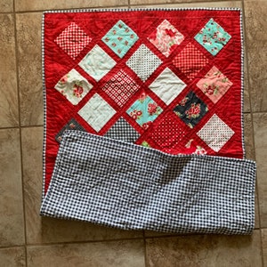 May include: A red and white quilt with a diamond pattern. The quilt is made of various fabrics with different patterns, including floral, polka dot, and plaid. The quilt is folded in half and has a black and white checked backing.