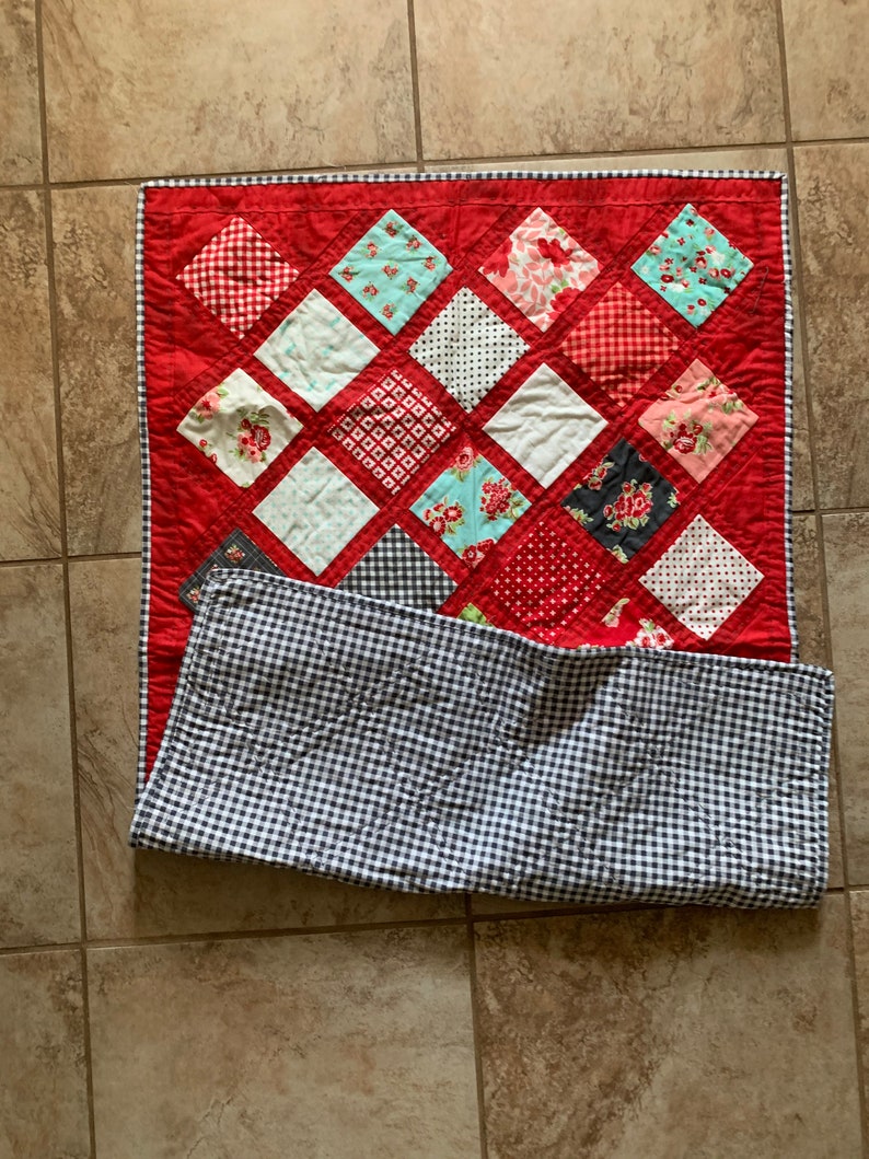 May include: A red and white quilt with a diamond pattern. The quilt is made of various fabrics with different patterns, including floral, polka dot, and plaid. The quilt is folded in half and has a black and white checked backing.