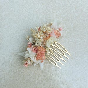 May include: A delicate hair comb featuring a cluster of dried flowers in shades of pink, cream, and beige. The floral arrangement is attached to a gold-toned metal comb, perfect for adding a touch of natural elegance to any hairstyle.