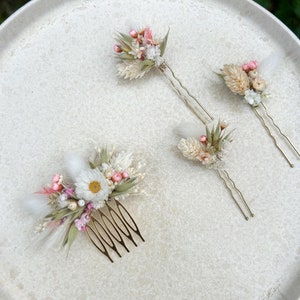 Peut inclure: Trois accessoires pour cheveux avec des fleurs séchées. Le premier est un peigne avec un bouquet de fleurs blanches et roses. Le deuxième et le troisième sont des épingles à cheveux avec des bouquets plus petits de fleurs blanches et roses.