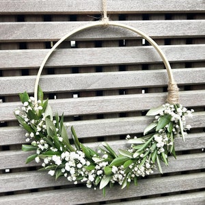 May include: A simple, rustic wreath made with white baby's breath flowers and green leaves, hanging on a wooden slat wall.