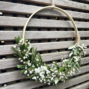 May include: A wooden hoop wreath decorated with white baby's breath flowers and green leaves. The wreath is hanging from a string on a wooden slat wall.