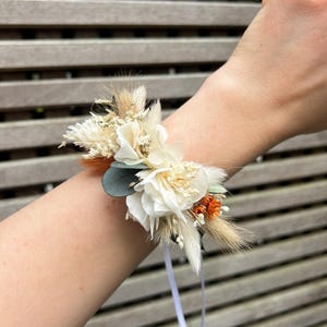 May include: A wrist corsage featuring a cluster of dried flowers in cream, orange, and green hues. The arrangement includes white blossoms, feathery grasses, and a small eucalyptus leaf, secured with a white ribbon.