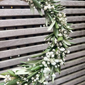 May include: A wreath made of green leaves and small white flowers. The wreath is attached to a wooden hoop.