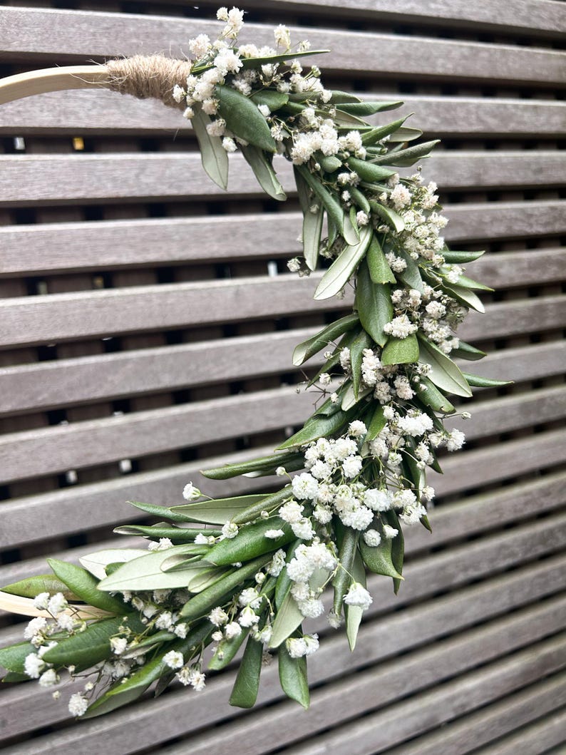 May include: A wreath made of green leaves and small white flowers. The wreath is attached to a wooden hoop.