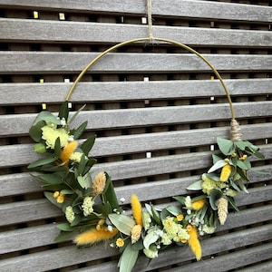 Könnte beinhalten: Ein dekorativer Blumenkranz mit einem goldenen Metallreifen. Er ist mit grünen Eukalyptusblättern, gelben flauschigen Blüten und kleinen weißen Blüten geschmückt. Er hängt an einem verwitterten Holzzaun, aufgehängt an einem Naturfaden.