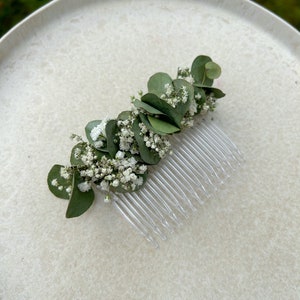 May include: A white and green floral hair comb with small white flowers and green leaves. The comb is made of clear plastic and has a row of teeth.