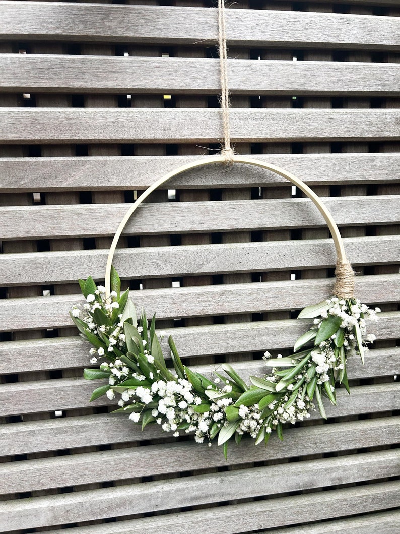 May include: A simple, rustic wreath made with white baby's breath flowers and green leaves, hanging on a wooden slat wall.