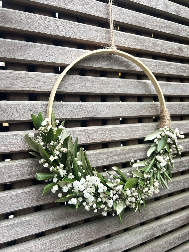 May include: A wooden hoop wreath decorated with white baby's breath flowers and green leaves. The wreath is hanging from a string on a wooden slat wall.