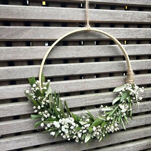 May include: A simple, rustic wreath made with white baby's breath flowers and green leaves, hanging from a wooden slat wall.
