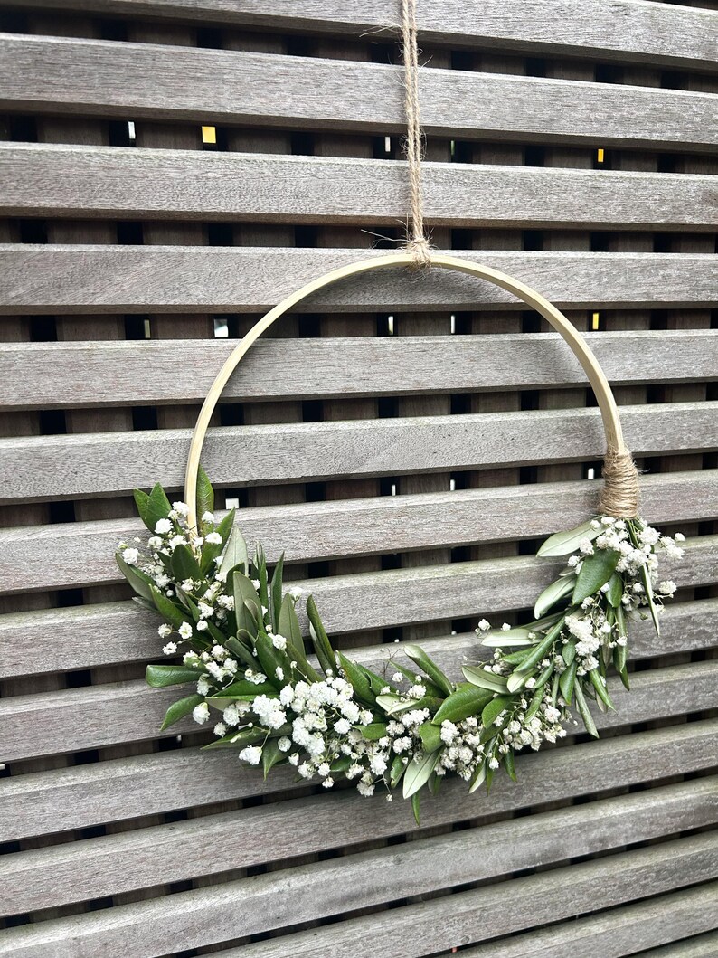 May include: A simple, rustic wreath made with white baby's breath flowers and green leaves, hanging from a wooden slat wall.