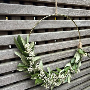 May include: A gold metal hoop wreath decorated with white baby's breath flowers and green leaves hanging on a wooden slat wall.