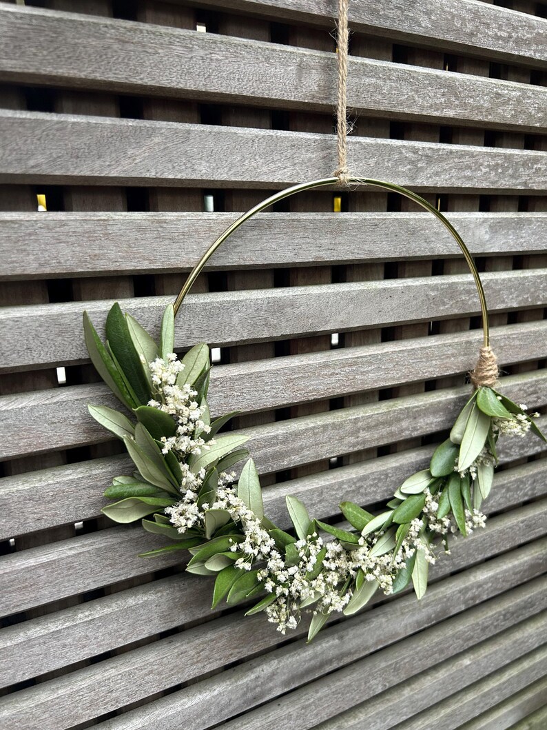 May include: A gold metal hoop wreath decorated with white baby's breath flowers and green leaves hanging on a wooden slat wall.