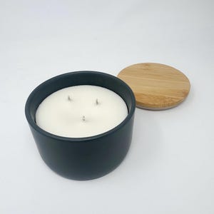 Puede incluir: Una vela azul oscuro en un recipiente de cerámica redondo con tres mechas. La vela tiene un relleno de cera blanca y una tapa redonda de bambú separada. La vela está sobre una superficie blanca.