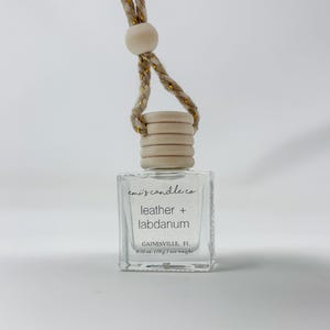 Può includere: Un profumatore per auto in vetro trasparente con tappo in legno e corda intrecciata. L'etichetta recita "emi's candle co" e "leather + labdanum". Il peso netto è di 10 g.