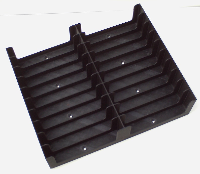May include: A black plastic tray with multiple vertical and horizontal dividers, creating a grid-like structure. The tray has a rectangular shape with raised edges and small circular holes. The dividers create individual compartments.