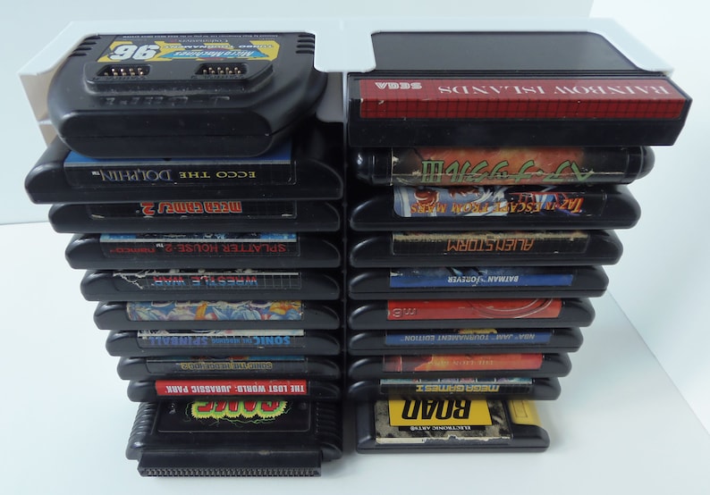 May include: A collection of black Sega Genesis video game cartridges, featuring titles such as "Sonic Spinball," "Ecco the Dolphin," and "Mega Games 2." The cartridges are held in a white holder.