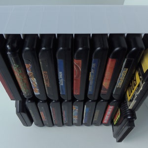 May include: A white plastic video game cartridge holder filled with black game cartridges. The cartridges have colourful labels with game titles. The holder is designed to keep the cartridges organised and accessible.