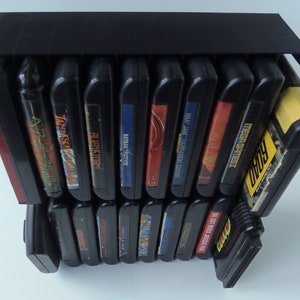 May include: A black video game cartridge storage unit, holding several game cartridges. The cartridges are black with colourful labels, featuring titles such as "Batman Forever" and "NBA Jam Tournament Edition". The unit is designed for display.
