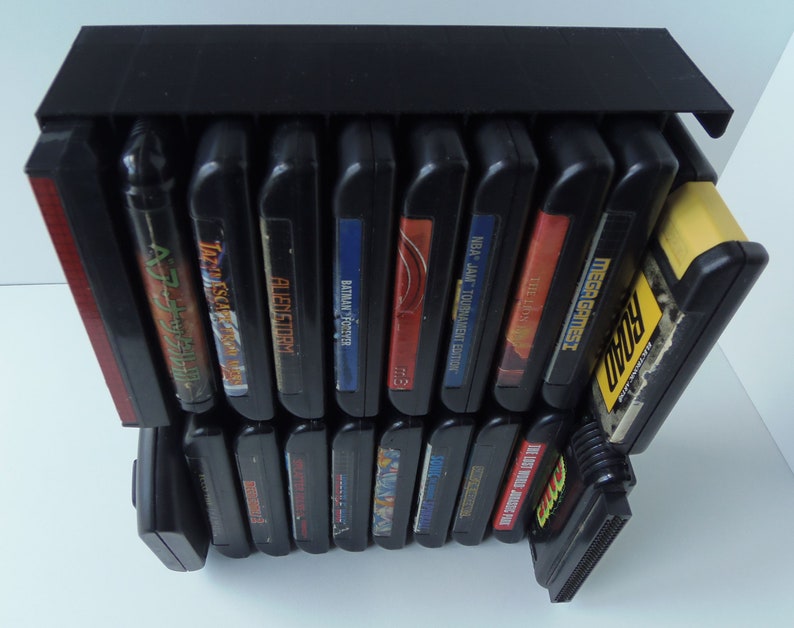 May include: A black video game cartridge storage unit, holding several game cartridges. The cartridges are black with colourful labels, featuring titles such as "Batman Forever" and "NBA Jam Tournament Edition". The unit is designed for display.