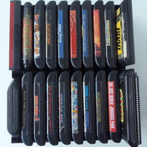 May include: A selection of black Sega Genesis video game cartridges in a black rack. Titles include "Alien Storm", "Batman Forever", "Sonic the Hedgehog 2", and "The Lost World: Jurassic Park".