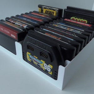 May include: A white plastic organiser filled with black video game cartridges. The cartridges have colourful labels with game titles like "Rainbow Islands" and "Micro Machines Turbo Tournament '96". The organiser is on a white surface.