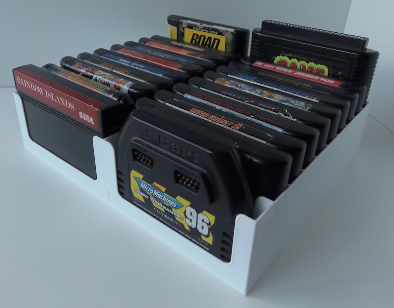 May include: A white plastic organiser filled with black video game cartridges. The cartridges have colourful labels with game titles like "Rainbow Islands" and "Micro Machines Turbo Tournament '96". The organiser is on a white surface.