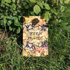 May include: A close-up of a small, yellow book with black ink illustrations and the words "Tick Magic" written on the cover. The book is lying on a bed of green grass.
