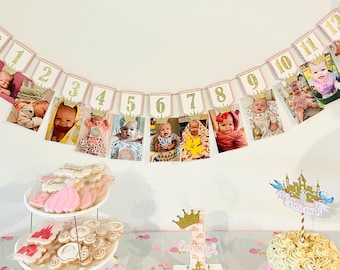 May include: A pink and gold banner with photos of a baby girl at each month of her first year. The banner is decorated with gold glitter crowns and numbers. The banner is hanging on a white wall.