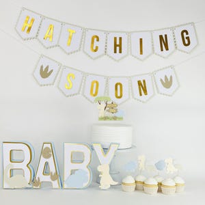 Può includere: Un set di decorazioni per baby shower a tema dinosauri. Include uno striscione con la scritta "HATCHING SOON", un cake topper e decorazioni per cupcake. La parola "BABY" è scritta in grandi lettere 3D. La combinazione di colori è bianco, oro e azzurro.