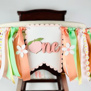 May include: Peach-themed high chair banner with peach, daisy, and ribbon decorations. The banner says "one" in pink letters.