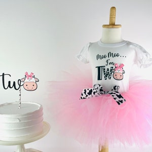 May include: A pink tutu with a black and white cow print ribbon. A white shirt with black text that says "Moo Moo... I'm Two" with a cow graphic. A white cake with a black and white cow cake topper that says "Two".