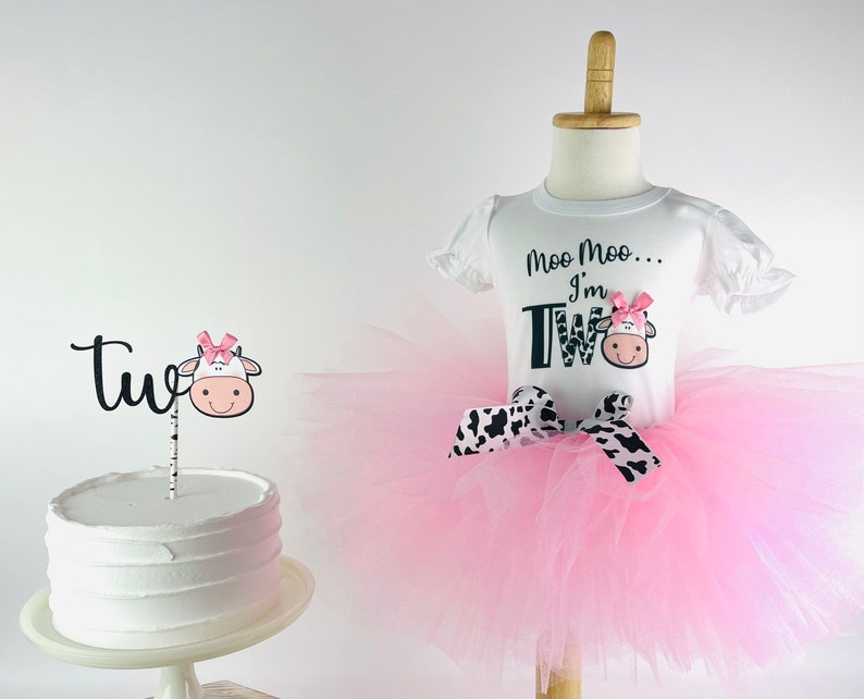 May include: A pink tutu with a black and white cow print ribbon. A white shirt with black text that says "Moo Moo... I'm Two" with a cow graphic. A white cake with a black and white cow cake topper that says "Two".