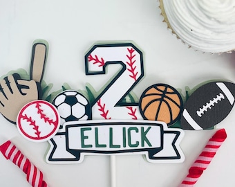 May include: A cake topper featuring a white number two with red stitching, a baseball, a football, a basketball, a baseball glove, a bat, and a banner with the name "ELLICK" in green letters.